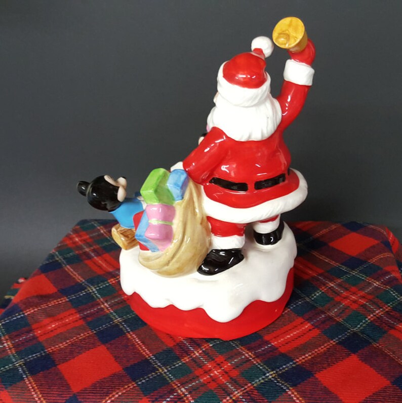 May include: A ceramic Christmas musical figurine of Santa Claus carrying a bag of presents with a small black and blue figure beside him. Santa is wearing a red suit with white trim and a black belt. He is holding a bell in his right hand and is looking to the right. The figurine is on a white base with a red bottom.