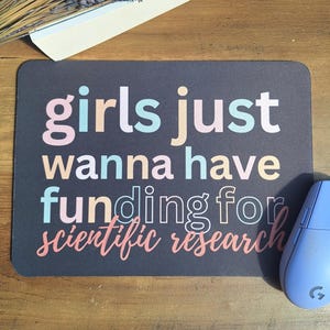 May include: A black mouse mat with the text "girls just wanna have funding for scientific research" in colourful letters.