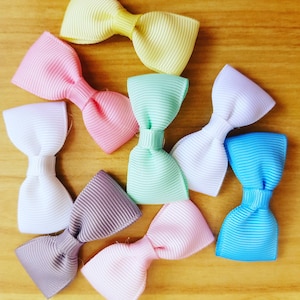 May include: Assortment of eight colorful ribbon bows in various colors, including pink, yellow, white, blue, green, and gray. The bows are arranged in a circular pattern on a wooden surface.