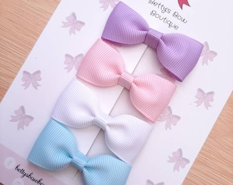 May include: Four pastel-colored hair bows in lavender, pink, white, and light blue are arranged on a white card with a pink bow logo and the text "Betty's Bow Boutique". The bows are made of ribbed fabric.
