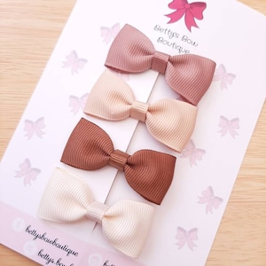 Puede incluir: Cuatro pequeños lazos para el pelo con textura acanalada en tonos malva, beige, marrón y crema están dispuestos sobre una tarjeta blanca. La tarjeta presenta el texto "Betty's Bow Boutique" y un logotipo de lazo rosa. Estos lazos son probablemente para niños.
