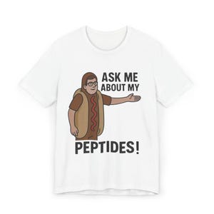 Puede incluir: Camiseta blanca con un personaje de dibujos animados disfrazado de perrito caliente, con gafas y una peluca marrón. El texto "ASK ME ABOUT MY PEPTIDES!" está impreso en negro debajo.