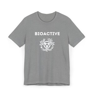 Puede incluir: Una camiseta gris jaspeado con la palabra "BIOACTIVE" en letras blancas sobre un gráfico blanco. El gráfico es un símbolo de peligro biológico estilizado con elementos florales.