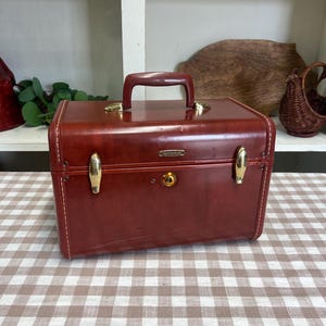 May include: A rectangular, dark red hard-shell vanity case with a matching top handle. The case features gold-tone metal latches, a central key lock, and a small gold-tone Samsonite logo plate on the front. The exterior has visible white stitching.