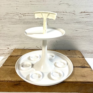May include: A white, three-tiered serving tray with a central support. The top tier has a handle, the middle tier is a flat circle, and the bottom tier has six circular indentations. The tray is made of a glossy, solid white material.