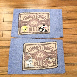 May include: Two blue denim pillowcases with brown and white cartoon characters. The pillowcases feature the text "Looney Tunes Quality Products Blues Trade Mark".