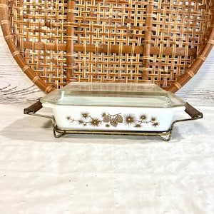 May include: A vintage white casserole dish with a clear glass lid, featuring a gold pinecone and branch design. It sits in a gold-toned metal rack with wooden handles. The dish is approximately 10 inches long.