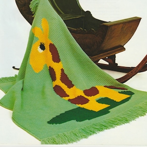 Puede incluir: Una manta de bebé verde tejida con un diseño de jirafa. La jirafa tiene la cabeza amarilla y manchas marrones. La manta tiene un borde con flecos y está colocada sobre una mecedora de madera.