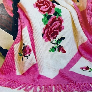 May include: A crocheted throw blanket with a pink and white diamond pattern and a floral design featuring pink roses. The blanket has a pink fringe border.