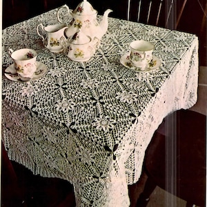 May include: A white crocheted tablecloth with a delicate floral pattern. The tablecloth is laid on a dark wooden table with a teapot, creamer, sugar bowl, and teacups.