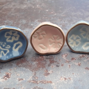 May include: Four small ceramic dishes with a gold rim and a white floral design on a blue, pink, and white background.