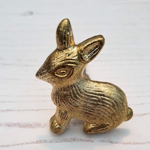 May include: A gold-coloured rabbit figurine with a detailed design. The rabbit is sitting on its hind legs with its front paws resting on the ground. The figurine is made of metal and has a shiny finish.