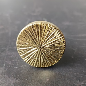 May include: A gold-coloured, circular cabinet knob with a textured surface. The knob features a radial pattern of lines radiating from the centre. The background is a dark, mottled surface.
