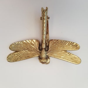 May include: A gold-toned metal dragonfly door knocker with intricate details. The wings are spread wide and the body is detailed with a segmented design.