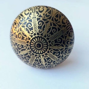May include: A round knob in black and gold with a decorative, intricate design. The knob features a raised, circular pattern in the centre.