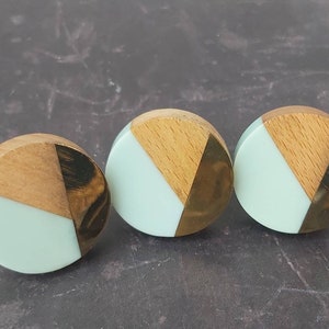 May include: Three decorative cabinet knobs with a geometric design. Each knob features a light green semi-circle, a wooden triangle, and a gold-coloured triangle. The knobs are round and appear to be made of wood, metal, and resin.