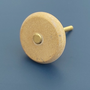 May include: A round, light brown cabinet knob with a gold screw and a gold metal centre. The knob is made of a textured material.