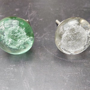 May include: Two glass cabinet knobs with silver metal bases. The knobs are clear glass with air bubbles inside. One knob is green and the other is clear.