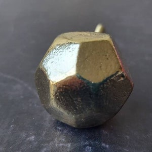 May include: A faceted, gold-coloured cabinet knob with a small, cylindrical stem. The knob has a geometric, multi-sided design, reflecting light. The hardware is likely for furniture, adding a decorative touch to drawers or doors.