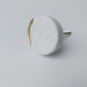 May include: White round marble cabinet knob with a gold metal accent. The knob has a screw on the back for attaching to a cabinet door.