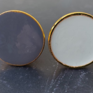 May include: Two round, decorative knobs with a gold rim. One knob is grey and the other is white.