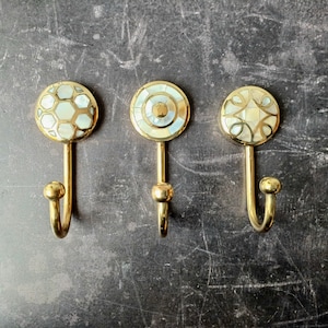 May include: Three gold-toned hooks with decorative white and gold inlays. The hooks are mounted on a grey surface.