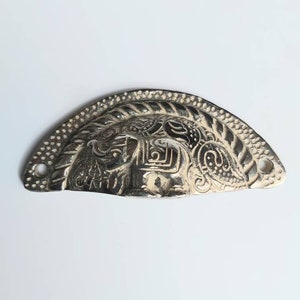 May include: Silver-toned metal cabinet pull with an ornate design featuring a stylised floral pattern. The pull is shaped like a half-circle and has two small holes for mounting.