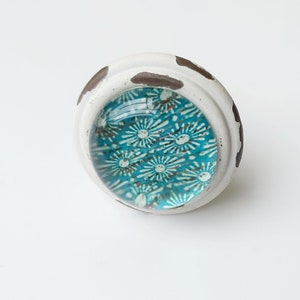 May include: A round cabinet knob in white and brown with a turquoise glass insert featuring a white floral pattern.