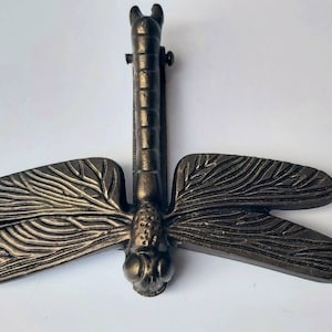 May include: Bronze dragonfly bottle stopper. The stopper has detailed wings with etched lines and a long, textured body. The dragonfly's head has large, round eyes. The stopper is designed to fit into a bottle.