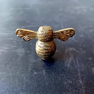 May include: A detailed, gold-coloured bee-shaped decorative object. The bee has textured wings and body, and is likely made of metal. The object is designed to be a decorative element, possibly for furniture or other craft projects.