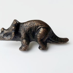 May include: A small, bronze-coloured dinosaur figurine in the shape of a Triceratops. The figurine is standing on a white surface.