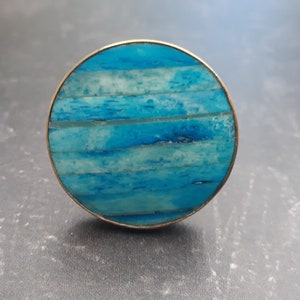 May include: A circular cabinet knob with a gold-coloured rim. The face of the knob has a mosaic design of blue and white rectangular pieces, forming a striped pattern. It is a decorative hardware element.