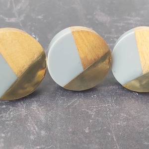 May include: Three round wooden knobs with a geometric design. The knobs are made of light grey and gold wood.