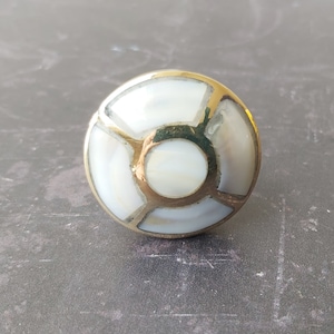 May include: A round, brass cabinet knob with a white mother-of-pearl inlay. The inlay is in a geometric pattern with a central circle.
