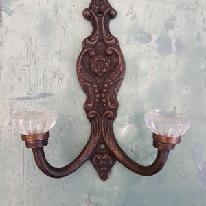 May include: An antique-style wall hook in a bronze finish, featuring two curved arms with clear glass knobs. The hook has an elaborate design with floral and scroll motifs, suitable for a vintage interior.