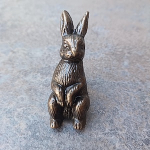 May include: A small, bronze-coloured rabbit figurine with a detailed, textured surface. The rabbit is sitting upright with its paws folded in front of it.