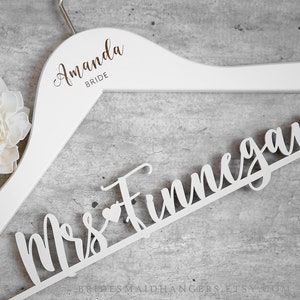 May include: White wooden hanger with the words "Amanda Bride" printed on the top and "Mrs.  Funnegan" in a decorative font on the bottom. A white flower is in the background.