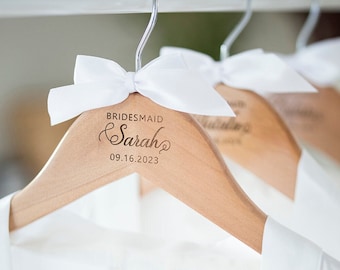 May include: Wooden hangers with white satin bows and the text "BRIDESMAID Sarah 09.16.2023" engraved on the front.
