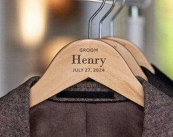 May include: Wooden hanger with the text "Groom Henry JULY 27, 2024" engraved on it. The hanger is hanging on a metal rod and has a brown blazer hanging on it.