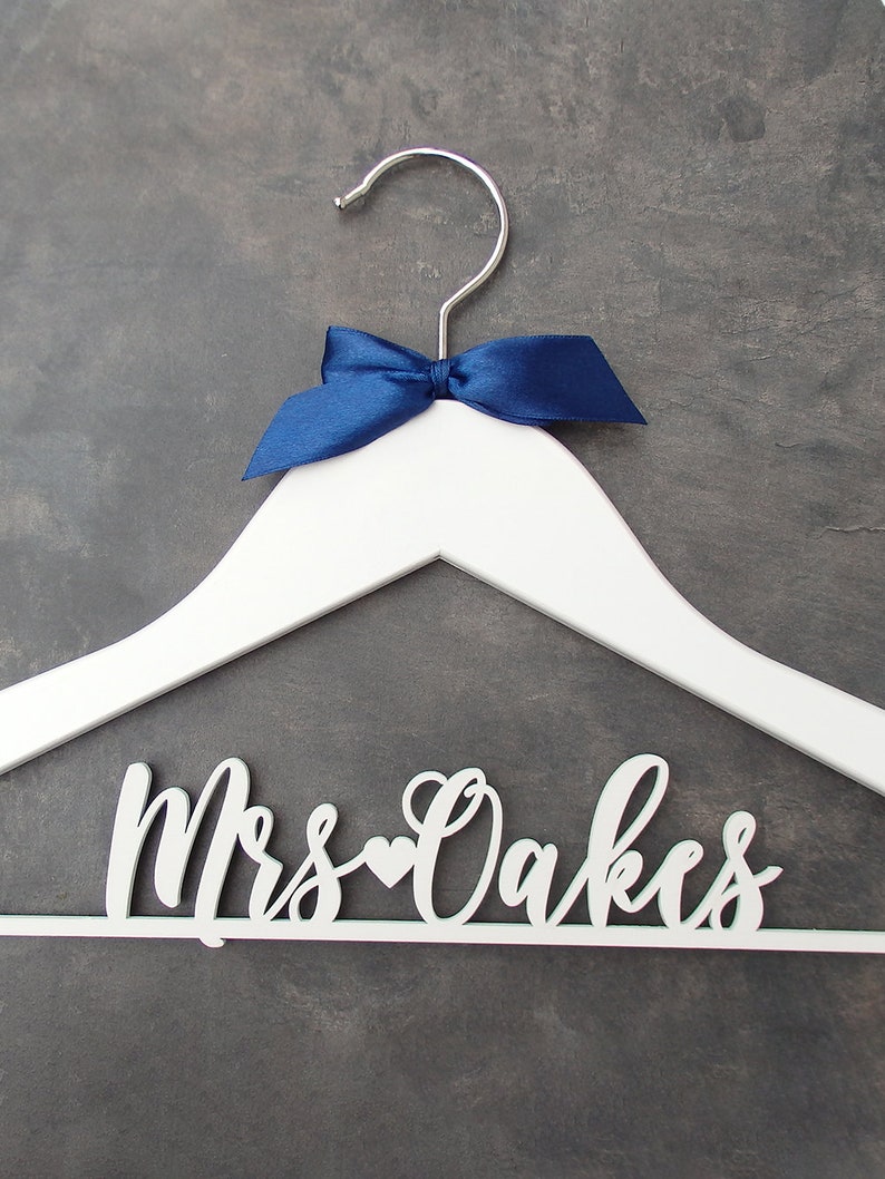 May include: White wooden hanger with a blue satin bow and the text "Mrs. Oakes" in white cursive lettering with a small heart between the words.