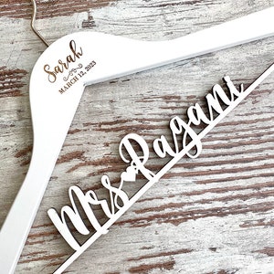 May include: White wooden hanger with the text "Sarah" and "March 12, 2023" on the top and "Mrs. Ragan" on the bottom.