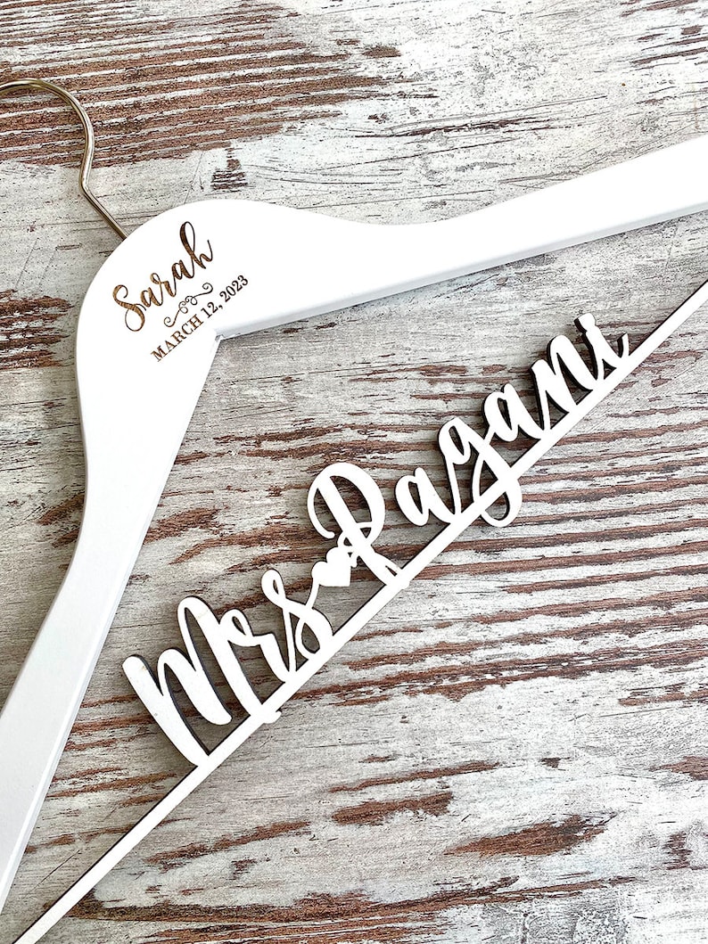 May include: White wooden hanger with the text "Sarah" and "March 12, 2023" on the top and "Mrs. Ragan" on the bottom.