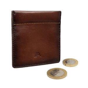 May include: A dark brown leather coin pouch with visible stitching along the top edge and a small embossed logo featuring the letter A with three vertical lines. Two metallic coins are positioned near the bottom right of the leather wallet.