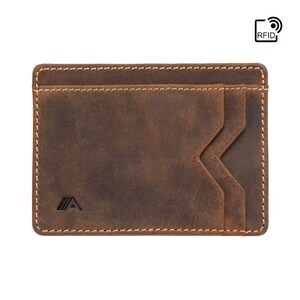 May include: A brown leather wallet with a stitched edge and a small logo on the front. The wallet has a flap that folds over the top and a pocket on the back. The wallet is RFID blocking.