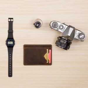 May include: A brown leather wallet with a card slot, a black digital watch with a white face, a silver film camera with a black lens, and a film canister on a light wood surface.