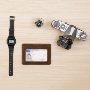 May include: A brown leather wallet with a clear plastic ID window, a black digital wristwatch with a white face, and a silver film camera with a black lens on a light wood surface.