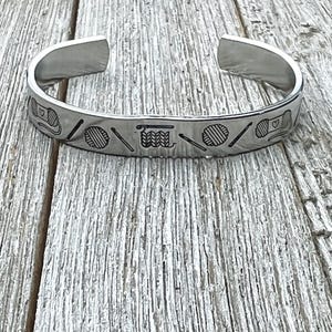 May include: A silver bangle bracelet with an engraved design of yarn balls, knitting needles, and a heart.
