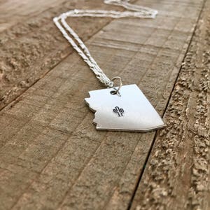 May include: A silver necklace with a pendant shaped like the state of Arizona. The pendant features a small cactus design.