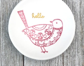 May include: A white ceramic dish with a pink bird design and the word "hello" in gold lettering.