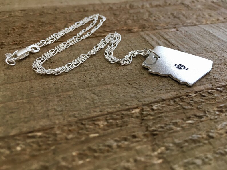 May include: Silver necklace with a pendant shaped like the state of Arizona featuring a small cactus design.